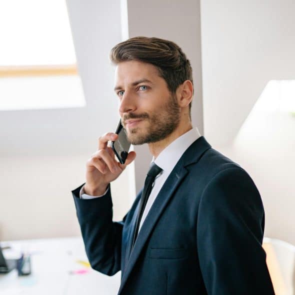 A man with light brown hair and a beard, wearing a navy suit and white shirt talking on a black smartphone in a bright, modern office setting.