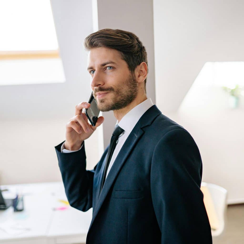 A man with light brown hair and a beard, wearing a navy suit and white shirt talking on a black smartphone in a bright, modern office setting.