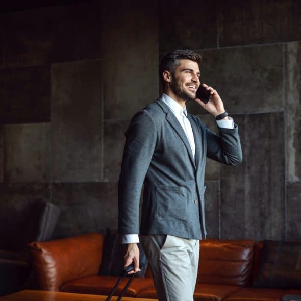 A man in a suit jacket talking on a cell phone and holding a suitcase.