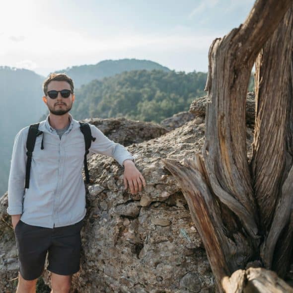 A man with a backpack standing on a rocky outcropping overlooking a mountainous landscape.