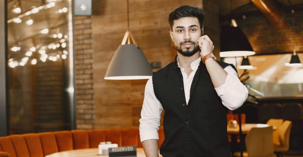 A man with dark hair and a beard is on the phone, wearing a black vest over a white long-sleeved shirt.