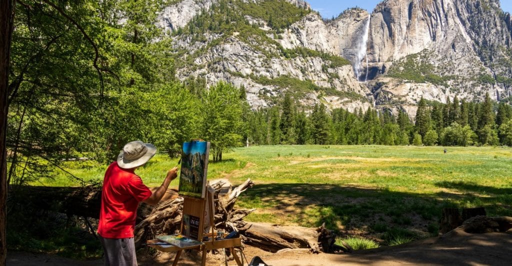 A man paints on a canvas outdoors with a mountain landscape backdrop.