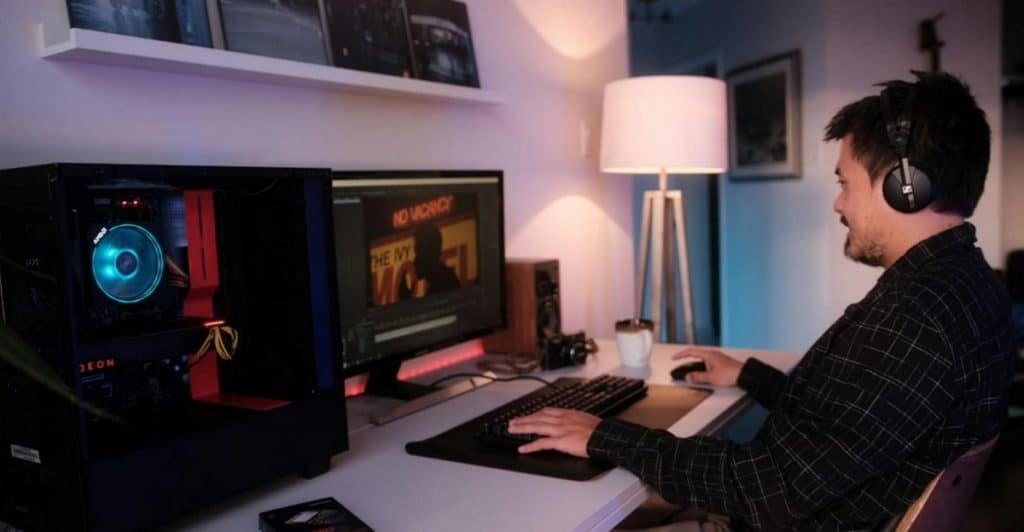 A man with headphones is sitting at a desk playing a computer game. The setup includes a desktop computer with a glowing fan, a monitor, a keyboard, and a mouse