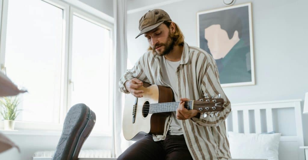 A man with long hair and a cap is playing a guitar and wearing a striped long-sleeved shirt.