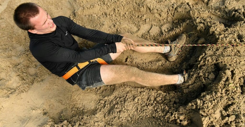 A short-haired man pulling a rope on an obstacle course in the sand.