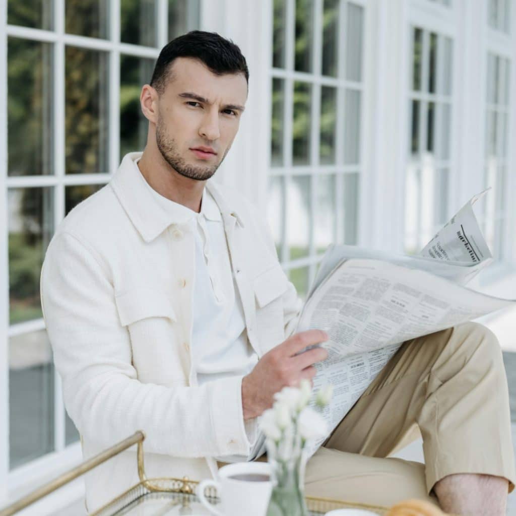 A man with short dark hair is sitting outdoors, reading a newspaper, and wearing a light-colored jacket over a white shirt and light-colored pants.