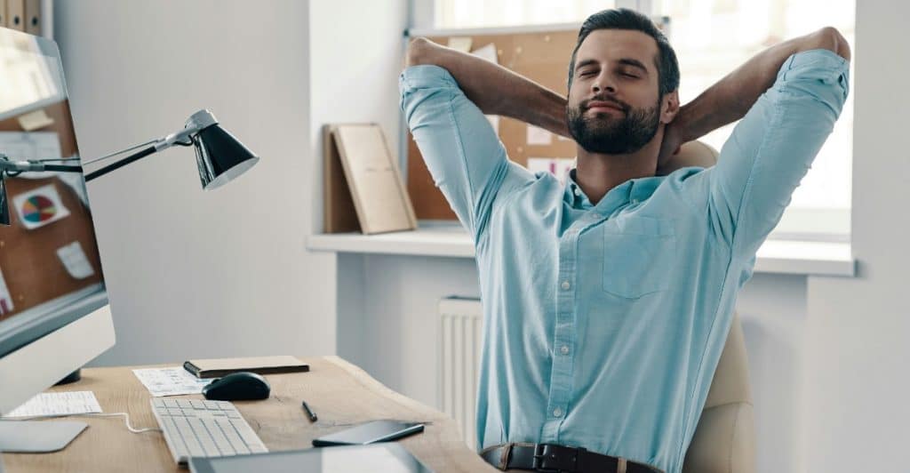 A man with dark hair and a beard, wearing a light blue shirt, leaning back in an office chair with his hands behind his head.