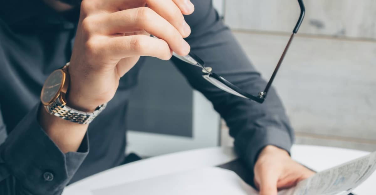 A man wearing a watch holding papers and eyeglasses, looking down at documents.
