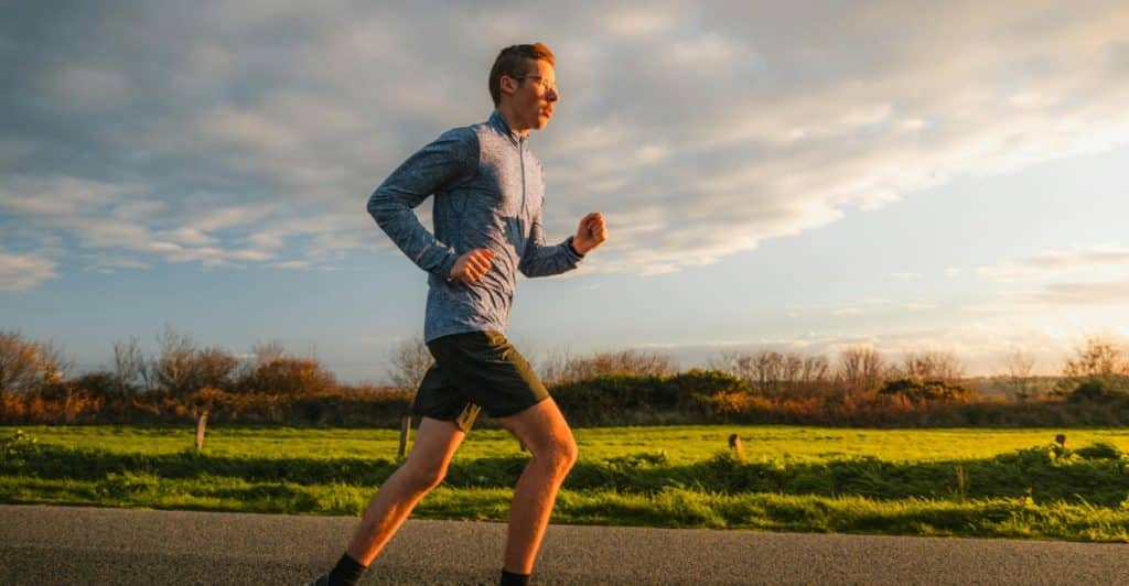 Man running on a road next to a field with the sun setting behind him.