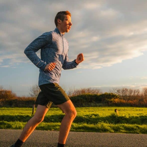 Man running on a road next to a field with the sun setting behind him.
