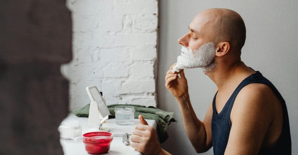 A bald man is applying shaving cream to his face while looking in a mirror.