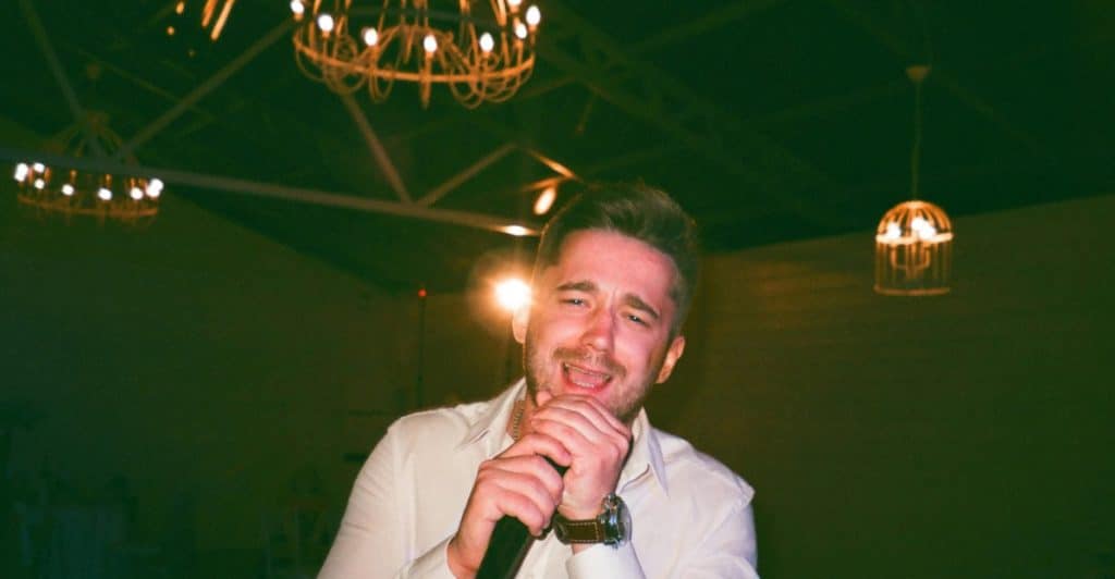 A man with a light beard holding a microphone and singing while wearing a white shirt in dim lighting with chandeliers in the background.