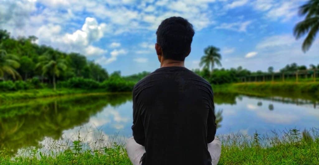 A person wearing dark clothing sitting on the grass facing a calm lake surrounded by greenery.