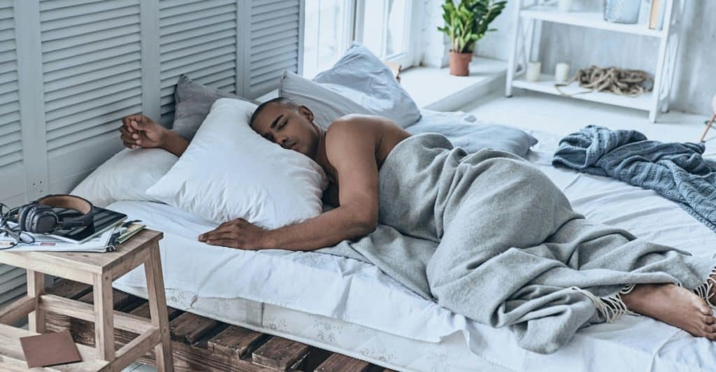 A man with short dark hair sleeping on his side in a bed with gray bedding.