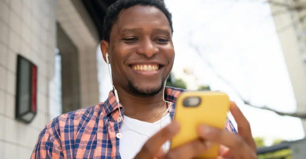 A man with wired earphones smiling as he looks at his yellow smartphone outdoors.