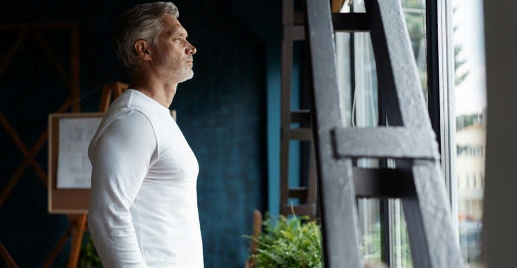A mature man with gray hair is standing and looking out a window.