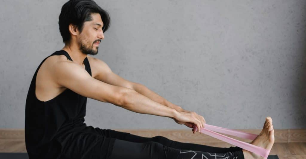 A man sitting on a yoga mat in a room with a concrete wall and wooden floor, stretching his arms forward and holding a pink resistance band with his feet, wearing a black tank top and black leggings with a white geometric design on the calf.