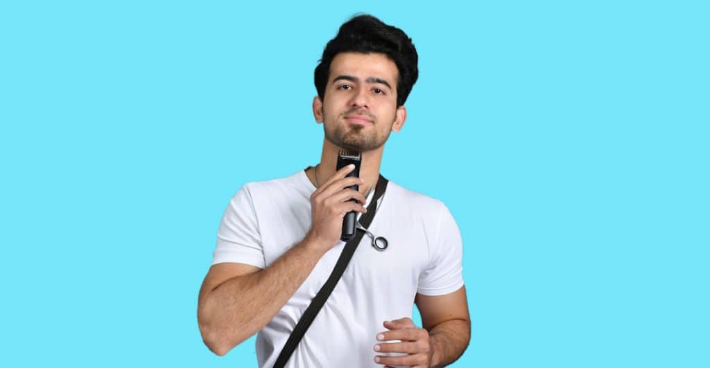 A man wearing a white t-shirt is using an electric trimmer to groom his facial hair against a bright blue background.