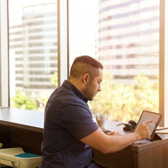 A man is sitting at a desk in front of a large window, using a tablet.