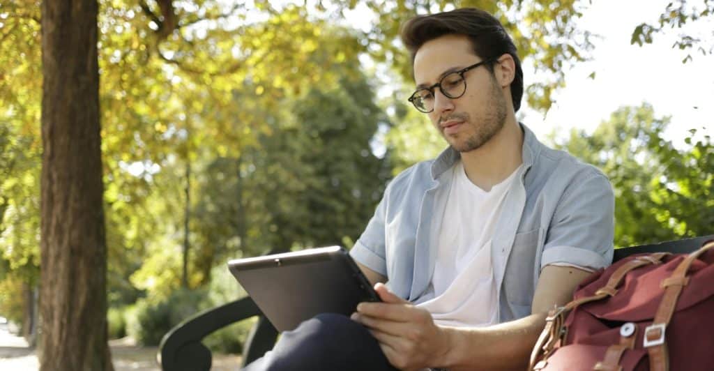 A man with glasses is sitting outdoors using a tablet and wearing a light blue short-sleeved shirt over a white t-shirt.