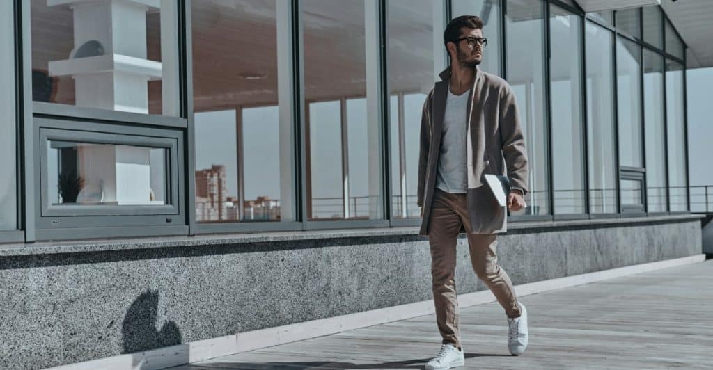 A stylish man wearing a coat and glasses is walking on a paved area in a city.