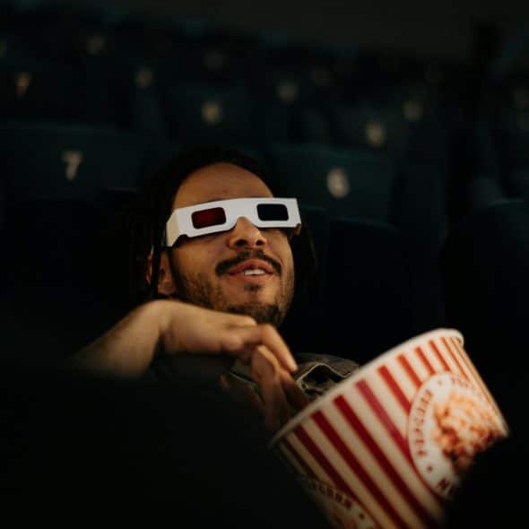 A man with dark hair and a beard wearing 3D glasses and holding a popcorn bucket in a movie theater.