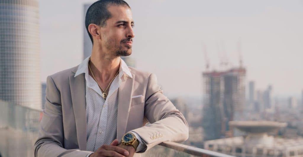 A man with short dark hair is wearing a light-colored blazer over a white shirt, with a city skyline in the background.