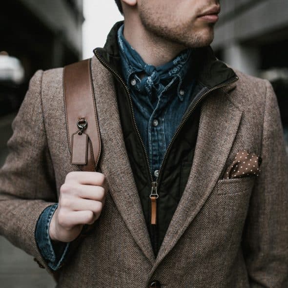 A man wearing a brown tweed jacket over a denim shirt and a dark vest, with a brown leather bag on his shoulder.