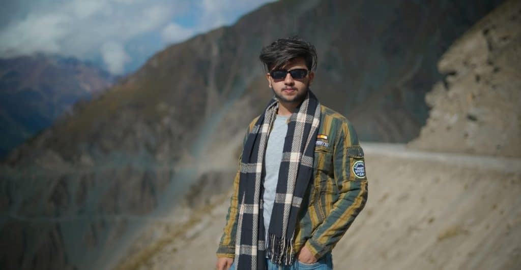 A man with dark hair and sunglasses is wearing a patterned scarf, a jacket, and jeans in a mountainous setting