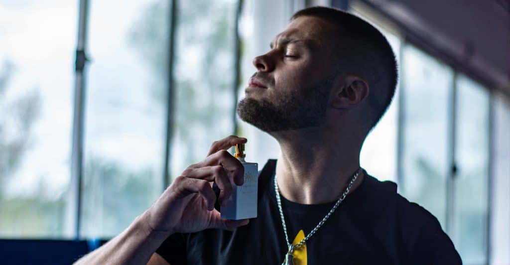 A man with short dark hair and a beard spraying cologne on his neck with windows in the background.