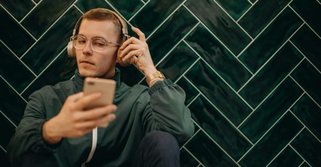 A man with glasses wearing a green jacket and headphones sitting against a green herringbone-patterned wall looking at his smartphone