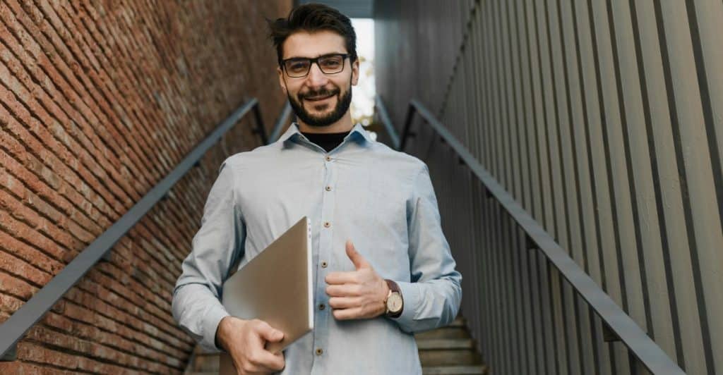 A man wearing glasses and a light blue shirt standing on a set of outdoor steps, holding a laptop and giving a thumbs-up.