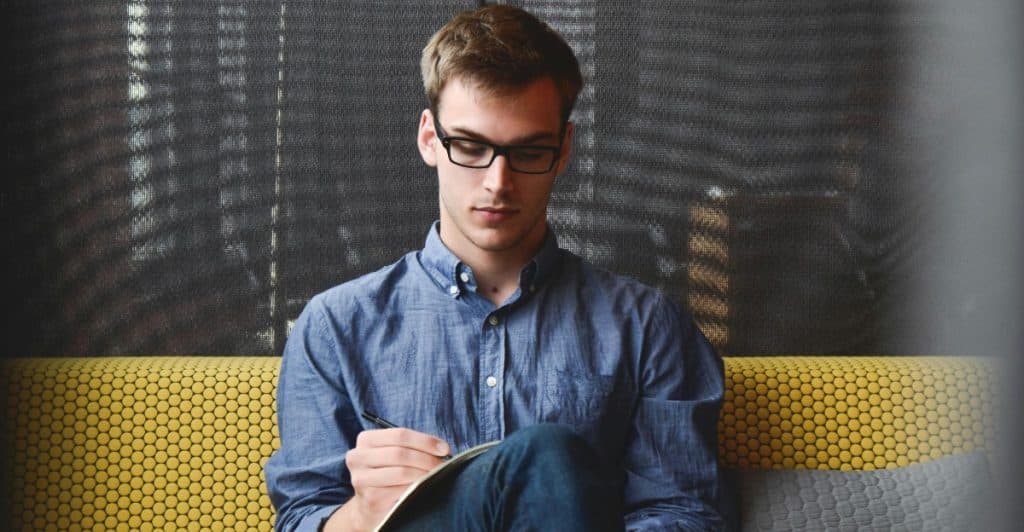 A man with glasses sitting on a yellow couch, writing in a notebook.