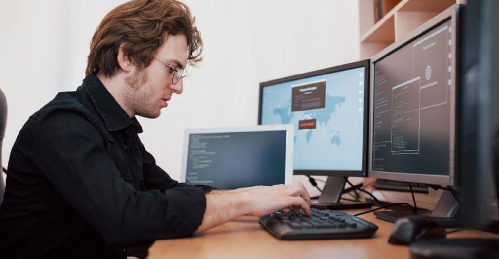 A man with reddish-brown hair and glasses typing on a keyboard in front of multiple computer monitors.