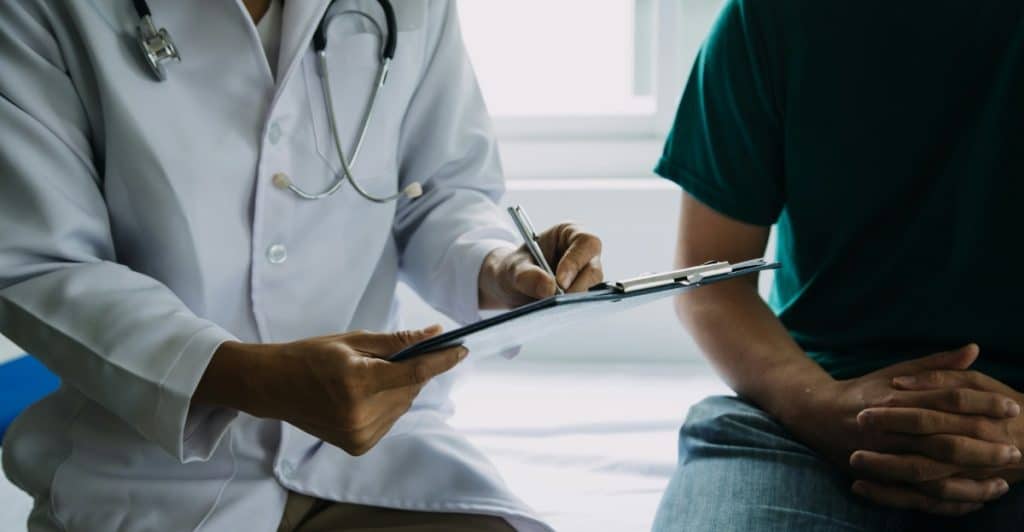 A medical professional in a lab coat is holding a clipboard and appears to be consulting with a patient.