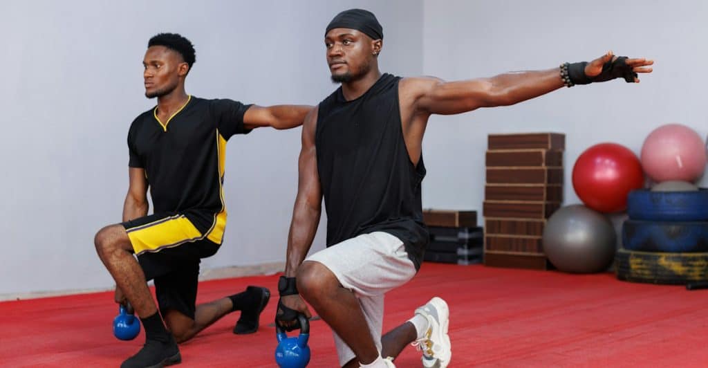 Two men with dark skin, wearing athletic wear, performing a kneeling lunge exercise with blue kettlebells in a gym with a red floor and various exercise equipment in the background.