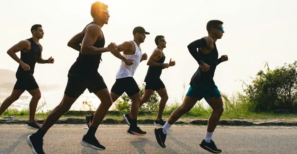 Five men against a bright sun, running together on a paved road, wearing athletic clothing and some with sunglasses.