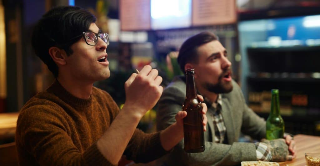 Two men sitting at a bar, eating chips and drinking from bottles, intently watching something off-screen.