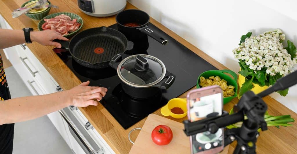An overhead view of a kitchen counter showing hands preparing food, pots on a stove, ingredients, and a phone set up on a tripod.