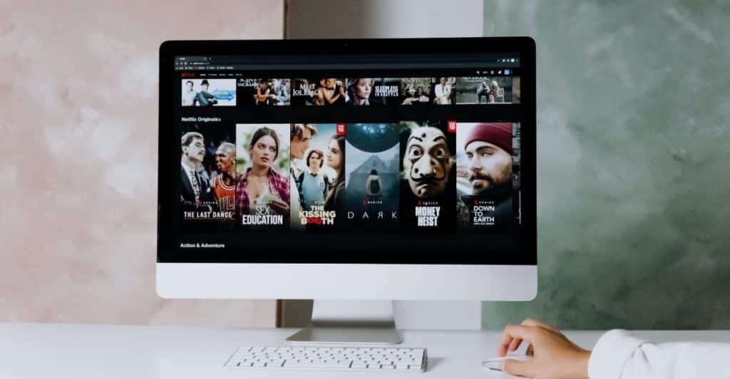 A computer monitor displaying the Netflix interface with various show and movie thumbnails is on a desk with a white keyboard and mouse.