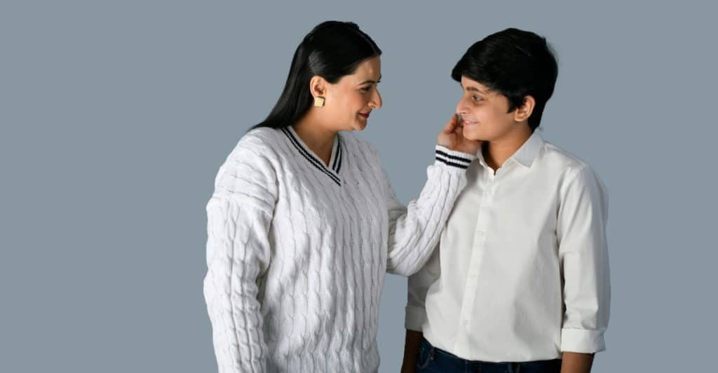 A mother placing her hand on the son's cheek leaning in to talk closely with him, with a neutral background.