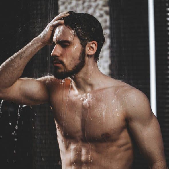 A person with wet hair and water droplets on their torso standing in a shower with dark tiles and bright vertical lights.