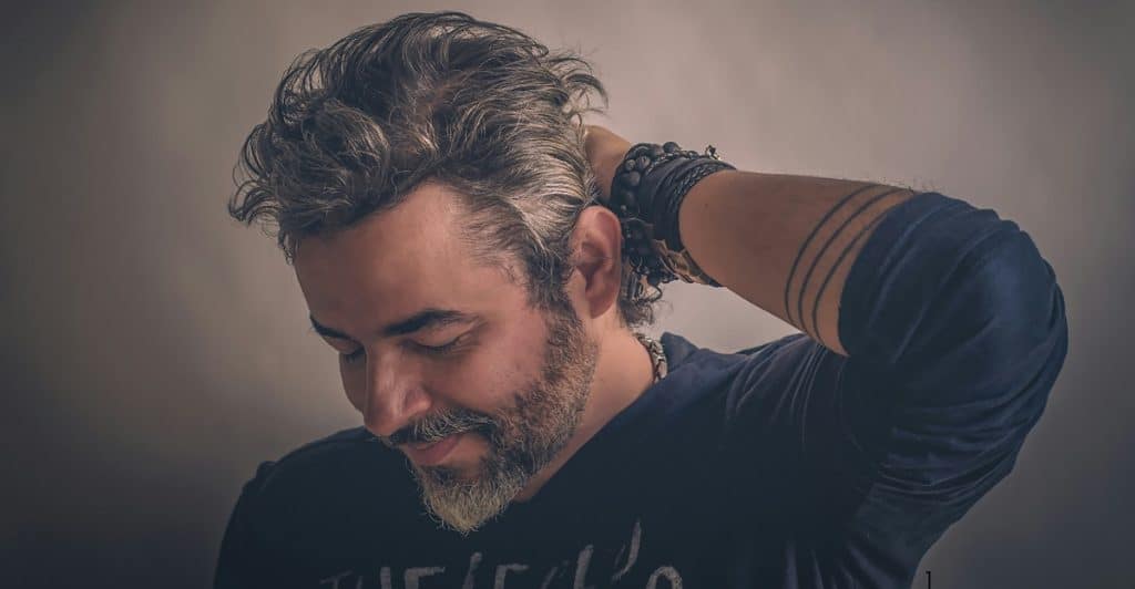 A man with styled hair and bracelets is looking down and to the side, with his hand near his neck.