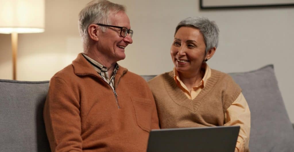 An older man and woman sitting close together on a grey couch, looking at each other and smiling warmly while a laptop rests on the man's lap.