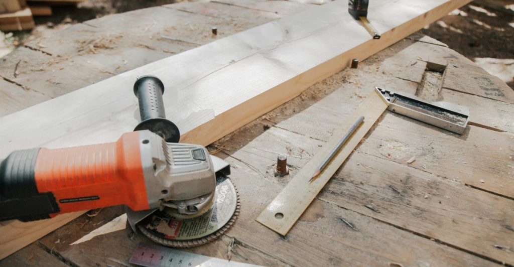 An orange angle grinder resting on a wooden surface next to a long wooden board, a ruler, and wood shavings.