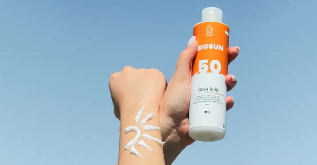 Hands holding a bottle of sunscreen and applying some to an arm in the shape of a sun against a blue sky.