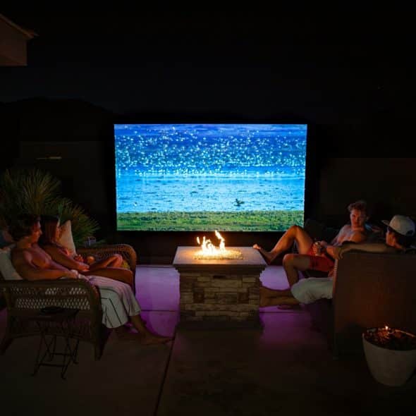 A group of people enjoying a movie on an outdoor screen at night, with a fire pit in the center.
