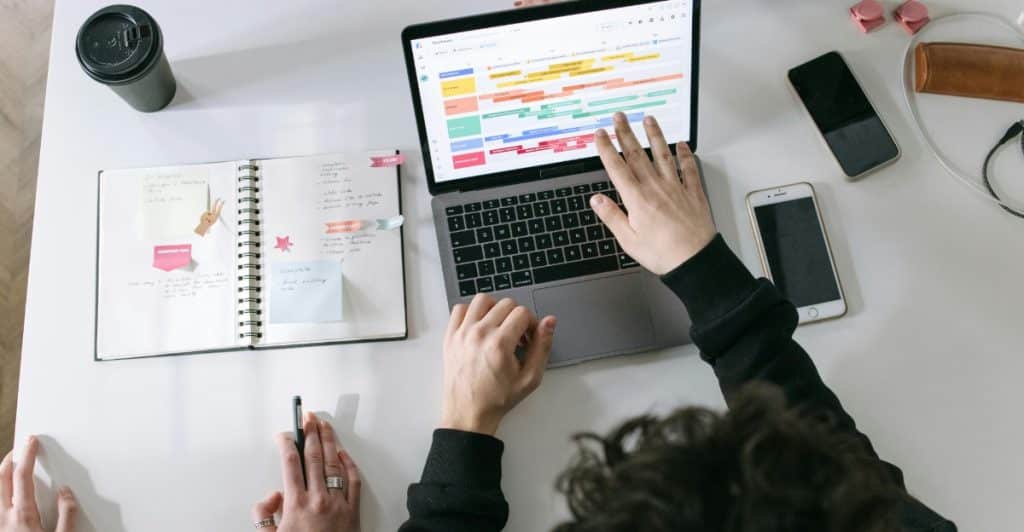 An overhead shot of two people's hands at a table with a laptop displaying a digital calendar and a physical notebook, showing organization and scheduling.