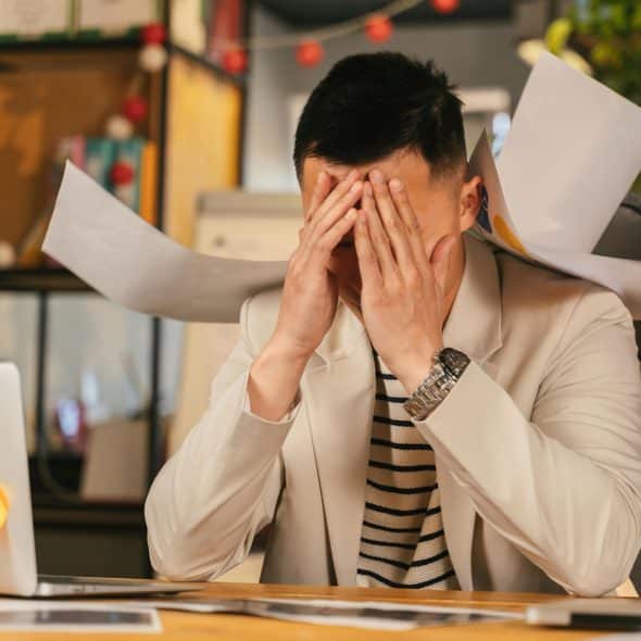 A person at a desk appears stressed, with hands covering their face and papers flying around.