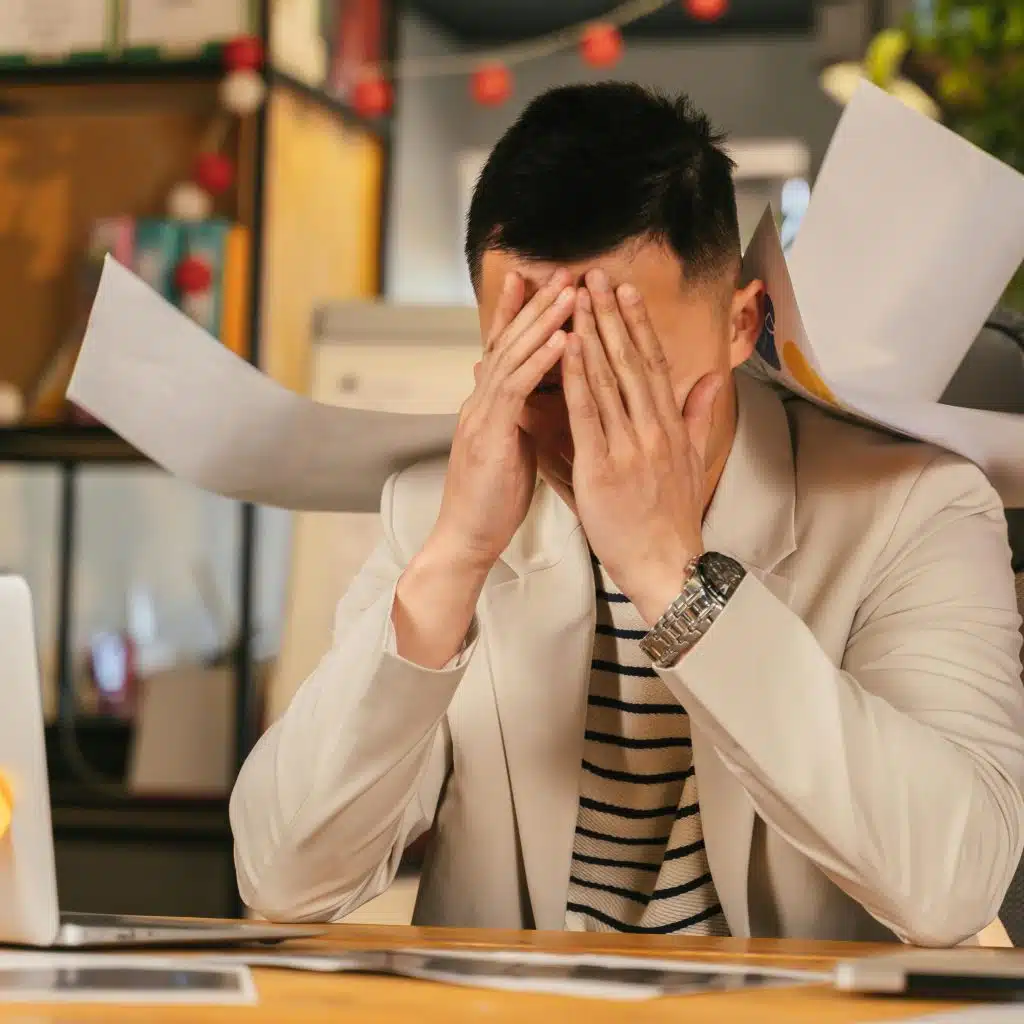 A person at a desk appears stressed, with hands covering their face and papers flying around.